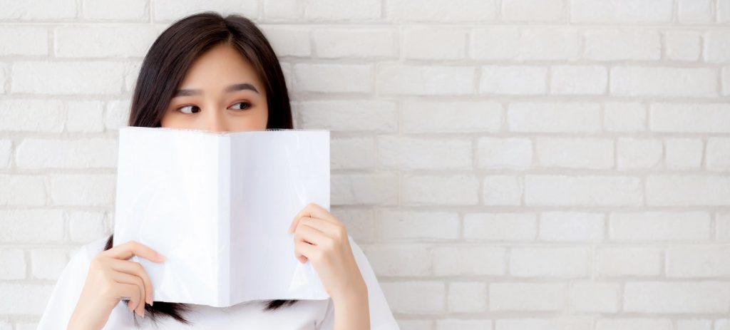 asian woman in front of a white brick wall hides her smile behind a book