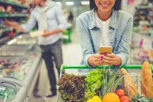 woman leaning on shopping cart full of vegetables in the store looking at her phone and smiling