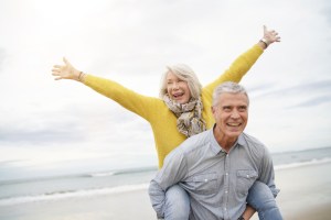 older couple on the beach with the wife in a yellow sweater piggybacking on her husband